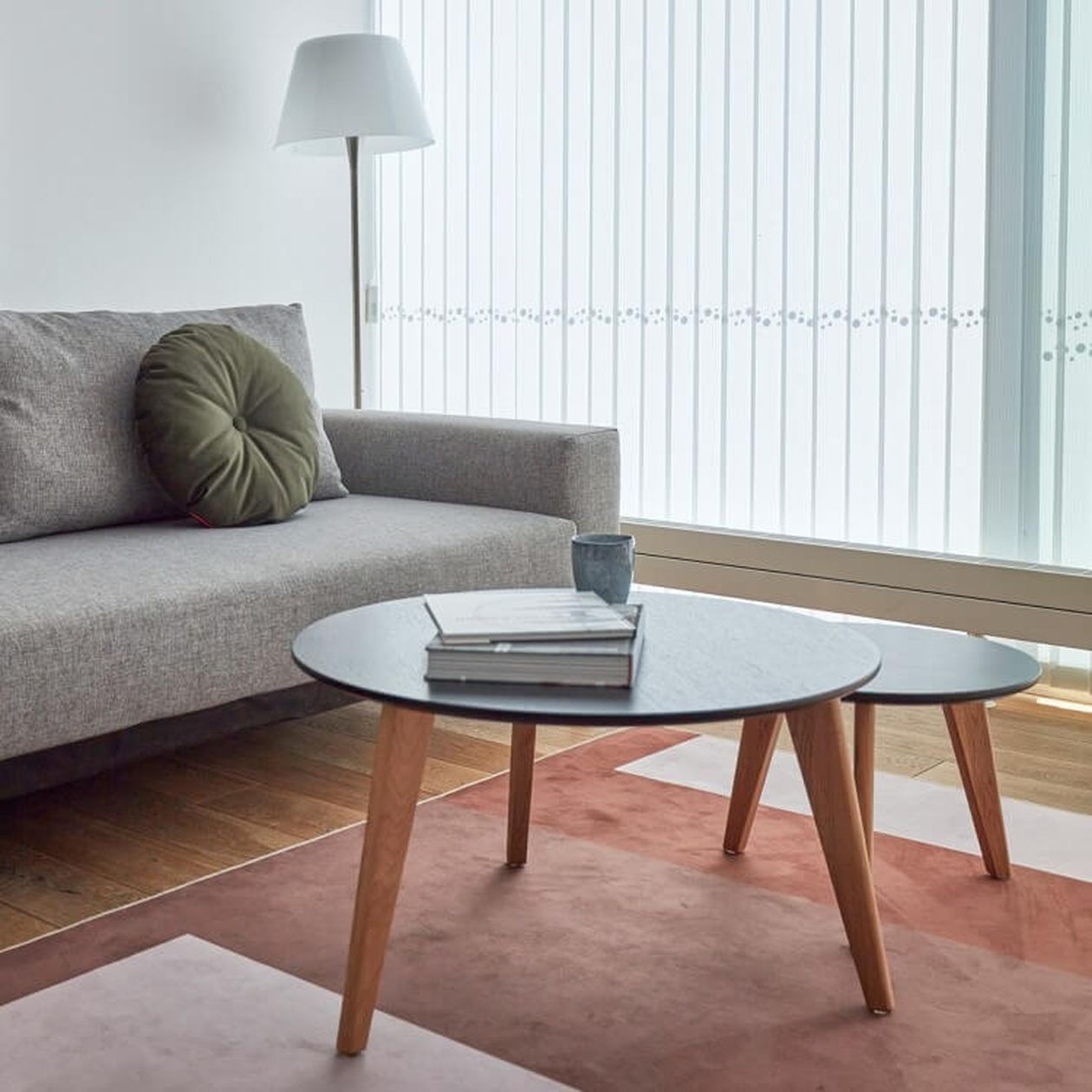 Moderne Wohnzimmereinrichtung mit einem grauen Sofa, einem runden Holztisch, Büchern, einer Tasse, einer Stehlampe und einem großen Fenster mit vertikalen Jalousien.
