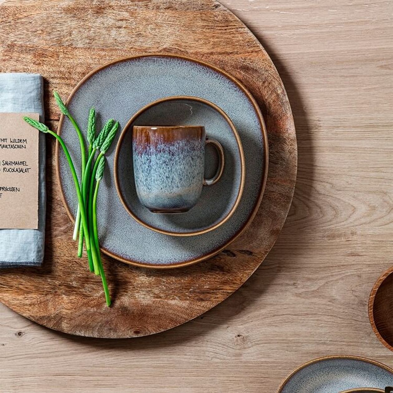Steingut-Geschirr in Blaugrau mit braunem Rand auf einem rustikalen Holzbrett, bestehend aus Tellern und Tasse mit Farbverlauf, dekoriert mit grünem Spargel.