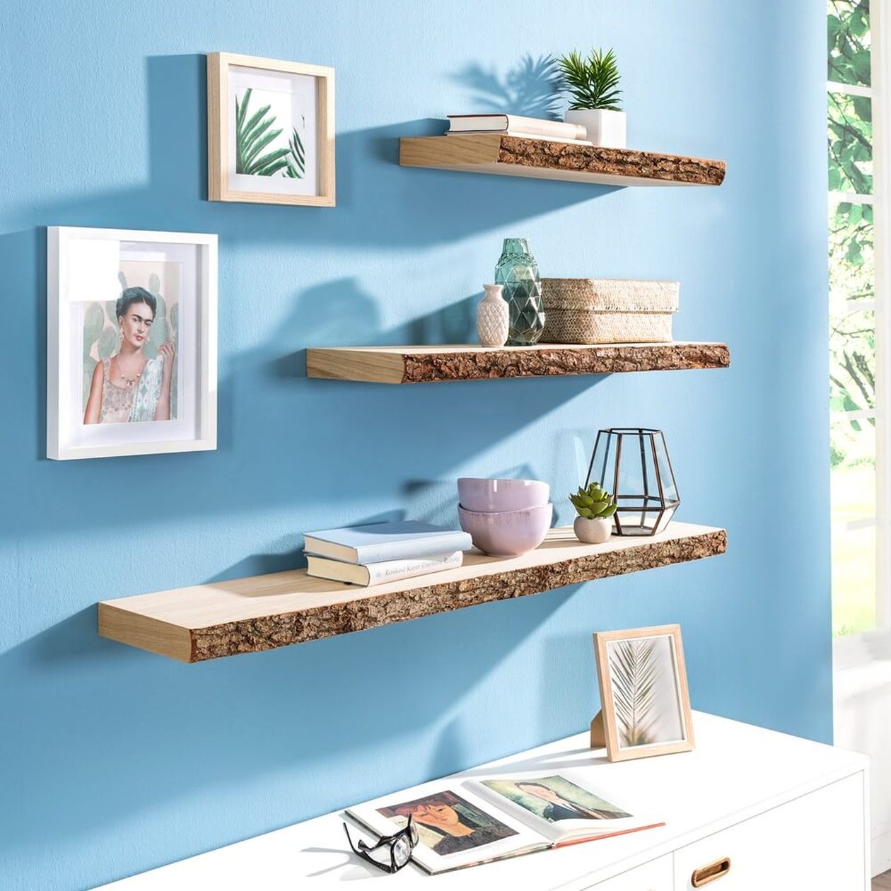 Holzregale mit Büchern, Pflanzen und Deko an einer blauen Wand. Gerahmte Kunst und ein Fenster mit Sonnenlicht runden die Raumdekoration ab.