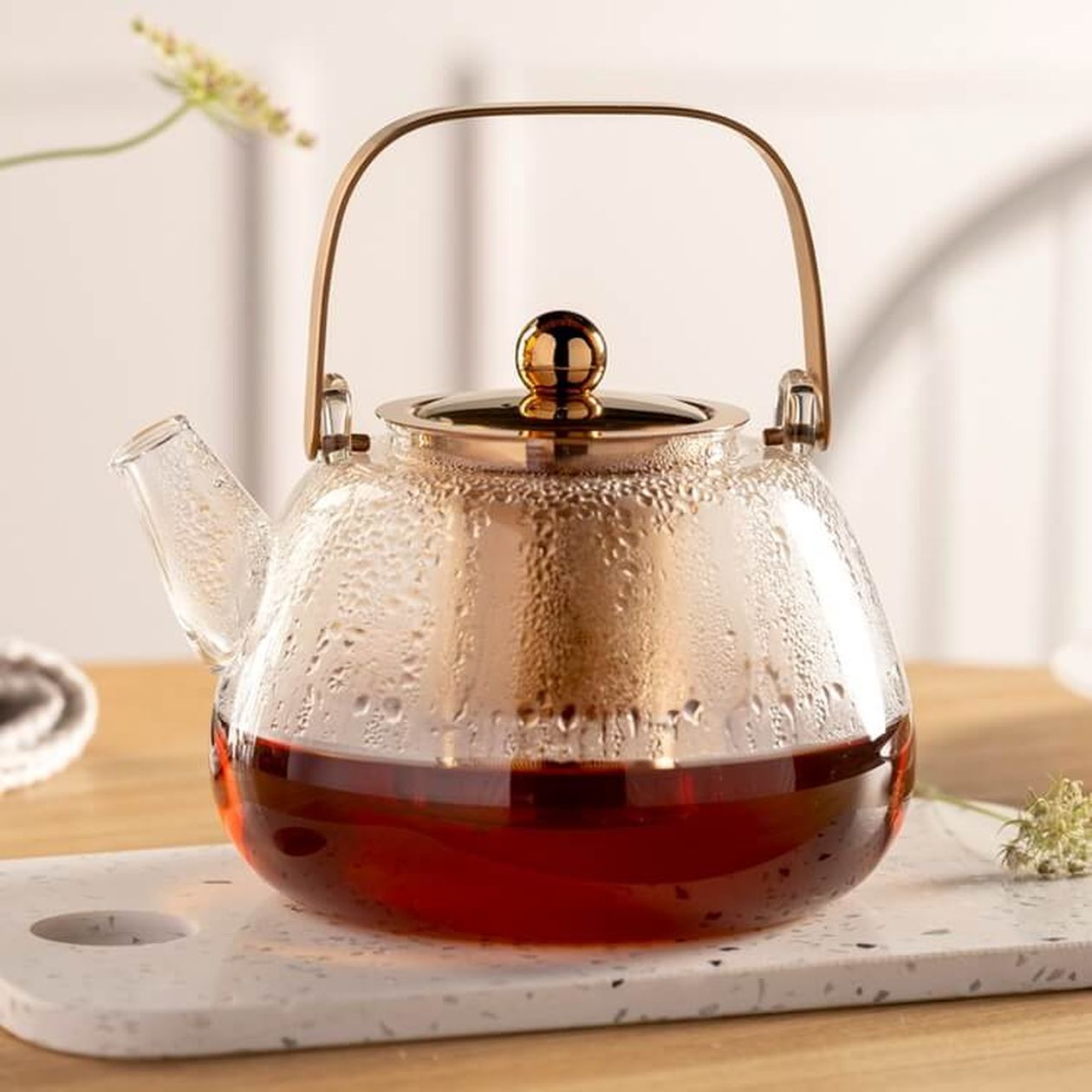 Bauchige Glasteekanne mit kupferfarbenem Deckel, Bügel und Einsatz, gefüllt mit rotem Tee, auf einem weißen Marmor-Tablett auf Holzuntergrund.