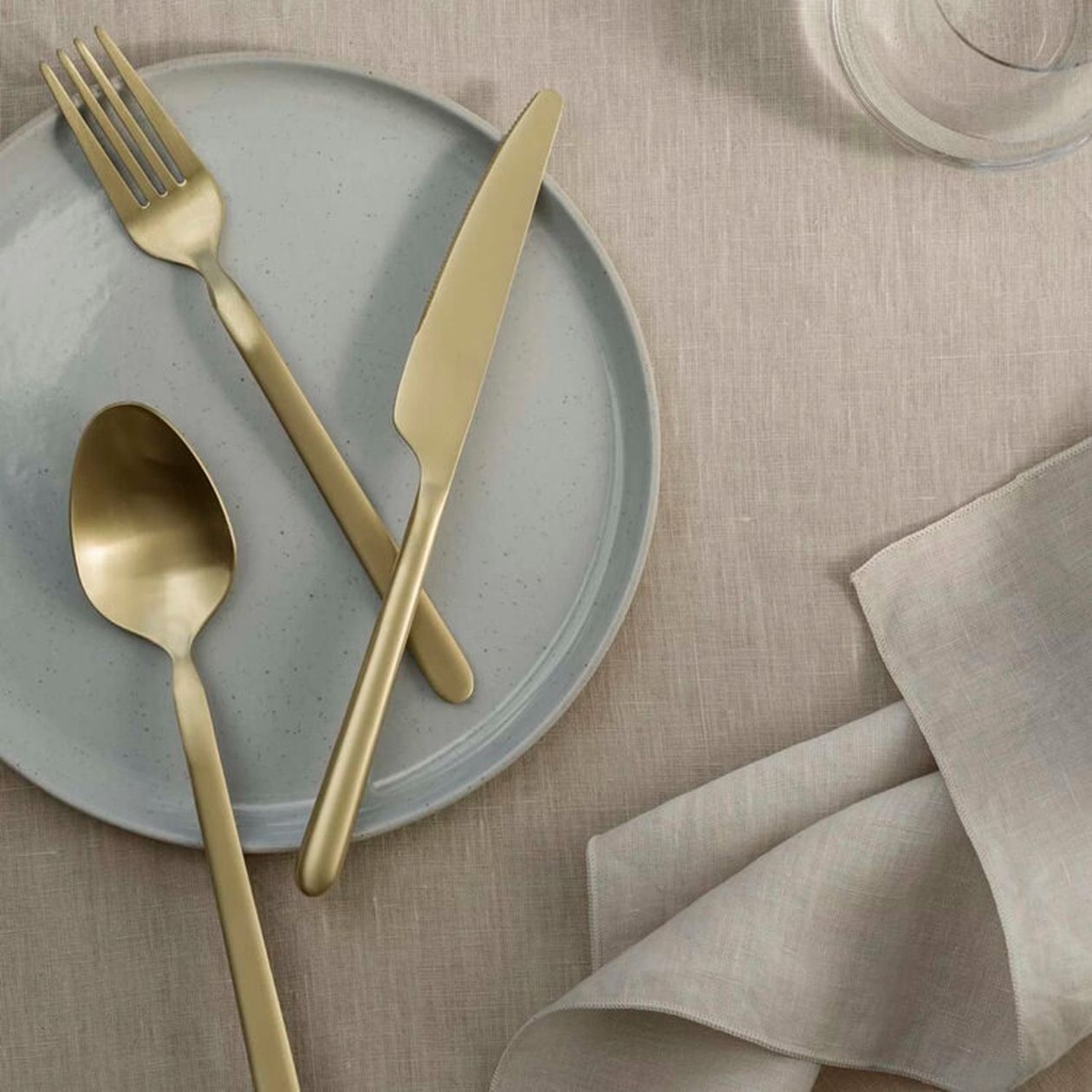 Mattgoldenes Besteck (Gabel, Messer, Löffel) auf einem hellgrauen Keramikteller, daneben eine naturfarbene Leinenserviette auf hellem Untergrund.
