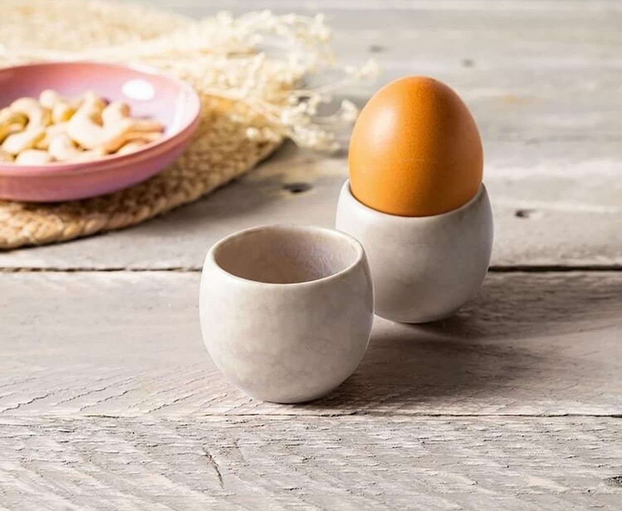 Zwei schlichte, bauchige Eierbecher aus Steingut in gebrochenem Weiß auf einem Holztisch, einer davon mit braunem Ei, im Hintergrund eine rosa Schale mit Cashewnüssen.