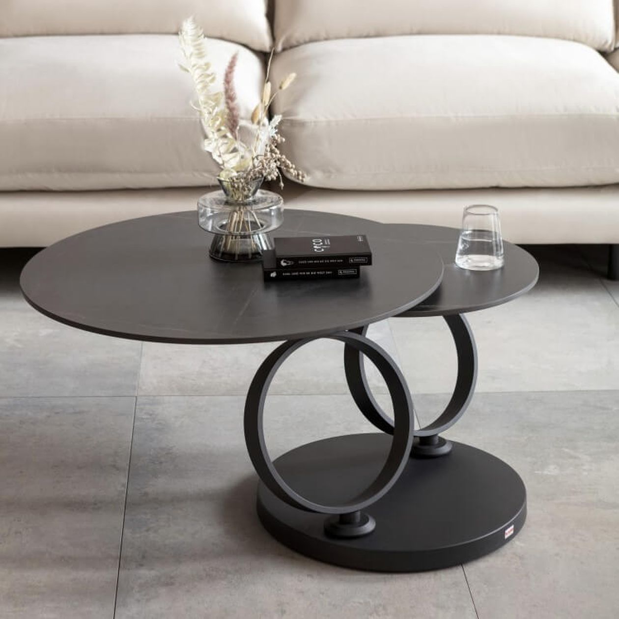 Moderner Couchtisch mit zwei runden schwarzen Flächen und kreisförmigen Stützen, auf dem ein Glas, Bücher und eine Vase stehen.