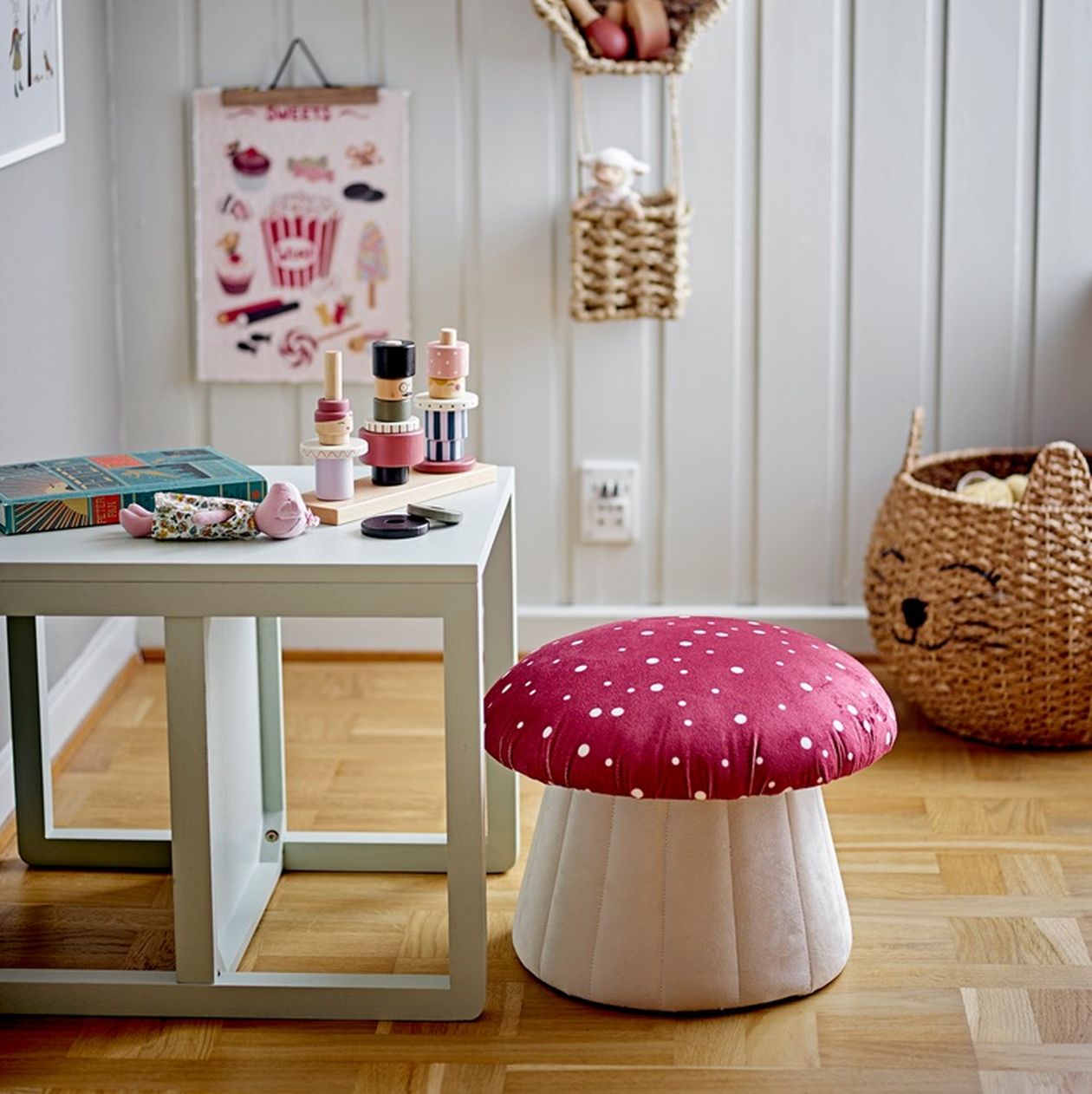 Kinderzimmer mit Hocker in Pilzform (weiß mit roten Punkten), daneben ein heller Spieltisch und Spielzeug, im Hintergrund ein Korb in Katzenform.