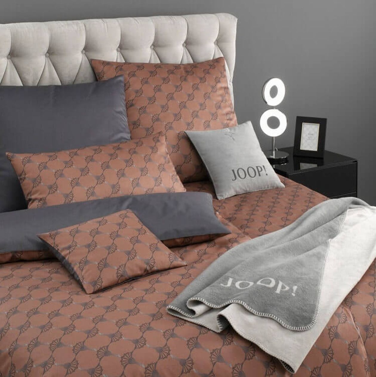 Elegantes Schlafzimmer mit gepolstertem Kopfteil, gemusterter braun-grauer Bettwäsche und Kissen. Über dem Bett liegt eine Decke mit der Aufschrift „JOOP!“.