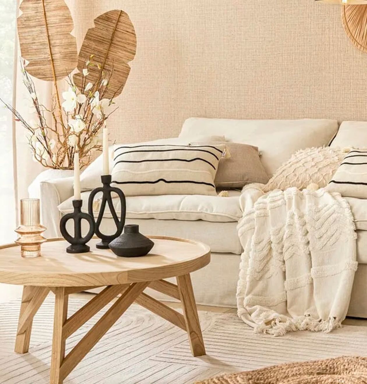 Runder Beistelltisch aus hellem Holz mit gekreuzten Beinen, dekoriert mit schwarzen Vasen und Kerzenhaltern, neben einem cremefarbenen Sofa in einem natürlich eingerichteten Wohnzimmer.
