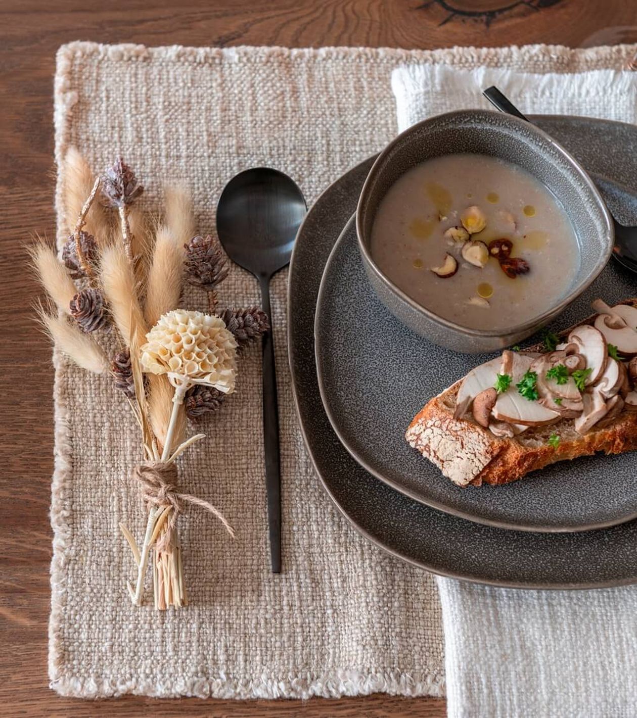 Eine Schüssel cremige Suppe mit Nüssen, serviert mit Brot mit Pilzen auf einer strukturierten Matte, neben einem rustikalen Blumenstrauß und einem schwarzen Löffel.