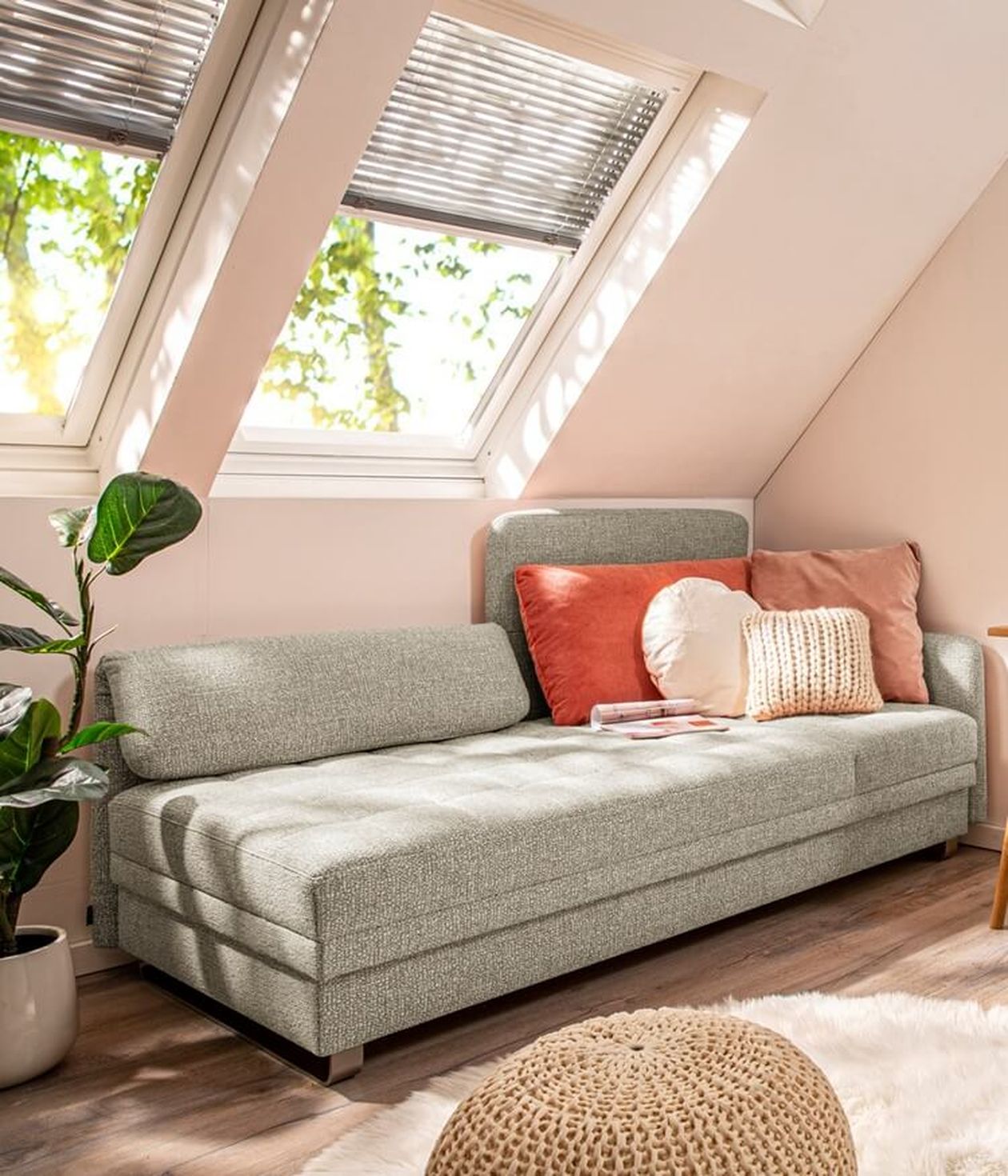 Gemütliches Dachzimmer mit hellgrauem Sofa, orange-weißen Kissen und Dachfenstern. Topfpflanze und Strickpouf auf dem Holzboden.