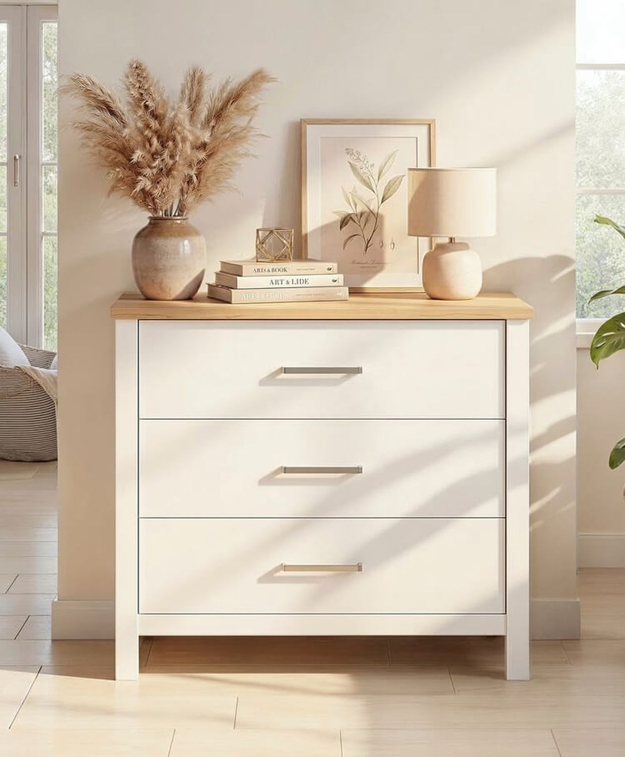 Weißes Sideboard mit heller Holzplatte und vier Schubladen mit silberfarbenen Griffen. Auf der Ablage stehen eine Vase mit Pampasgras, gestapelte Bücher, ein kleines goldenes Deko-Objekt, ein gerahmtes Bild mit Pflanzenmotiv und eine beigefarbene Tischlampe. Das Möbelstück steht in einem hellen, sonnendurchfluteten Raum mit hellem Holzboden.