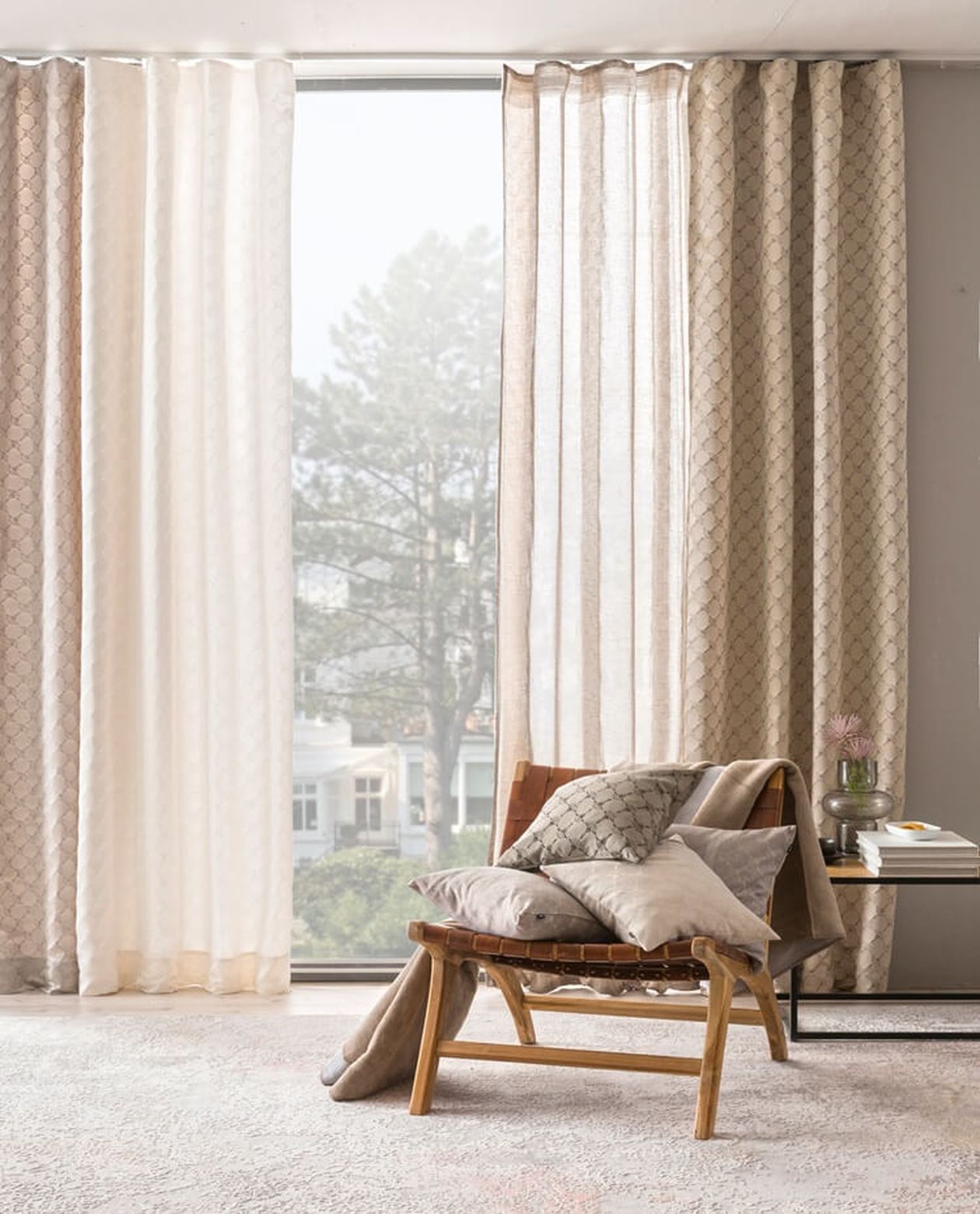 Helle, halbtransparente Fertiggardinen in Weiß und Beige mit dezentem Rautenmuster vor einem bodentiefen Fenster. Davor steht ein moderner Sessel mit Holzgestell und Ledergeflecht, dekoriert mit gemusterten Kissen in Grau- und Beigetönen. Rechts daneben ein kleiner Beistelltisch mit Büchern und einer Vase. Durch das Fenster ist ein Garten mit Bäumen sichtbar.