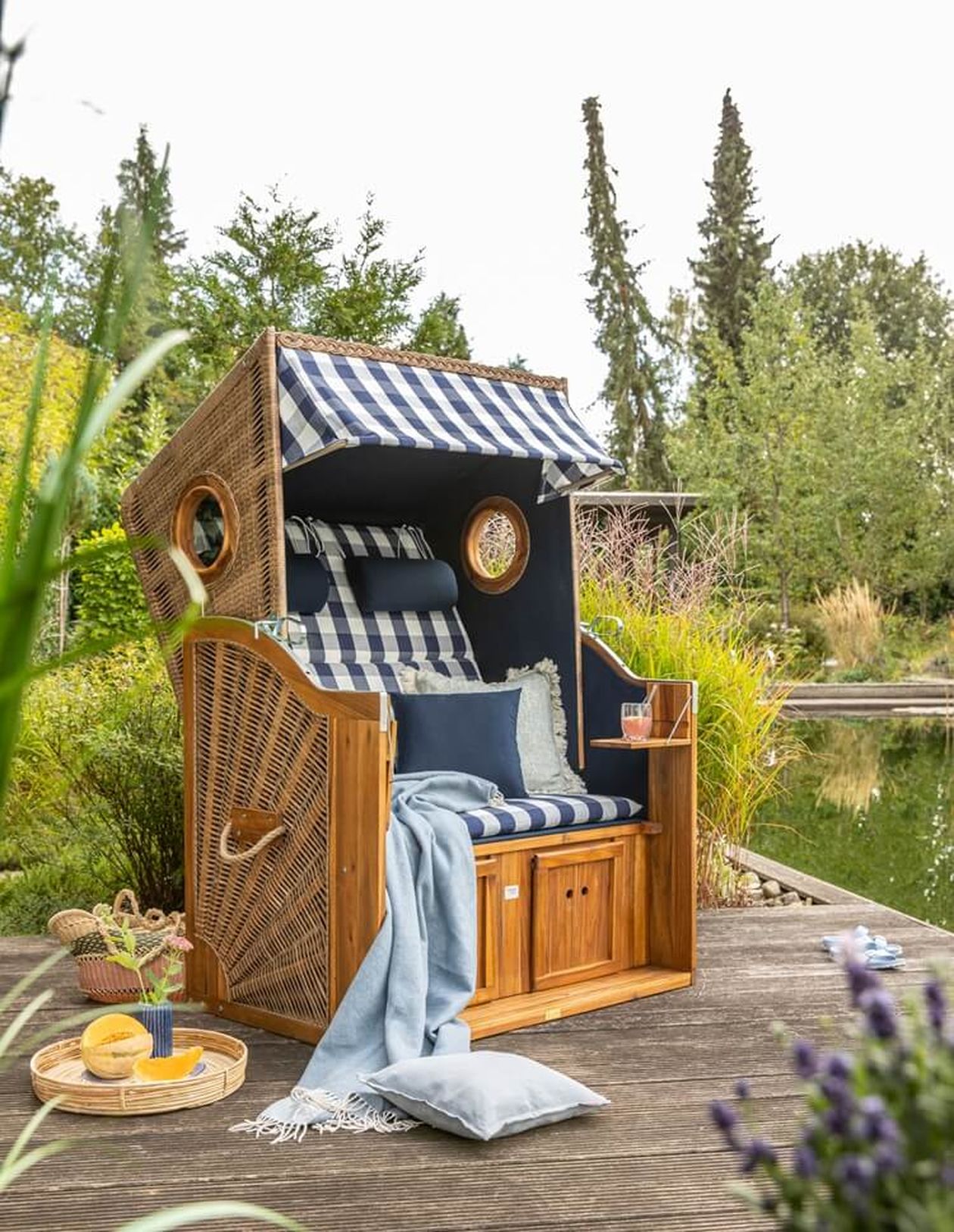 Klassischer Strandkorb aus Rattan mit blau-weiß karierter Polsterung auf einer Holzterrasse am Teich. Dekoriert mit Kissen, Decke und Tablett mit Getränken.