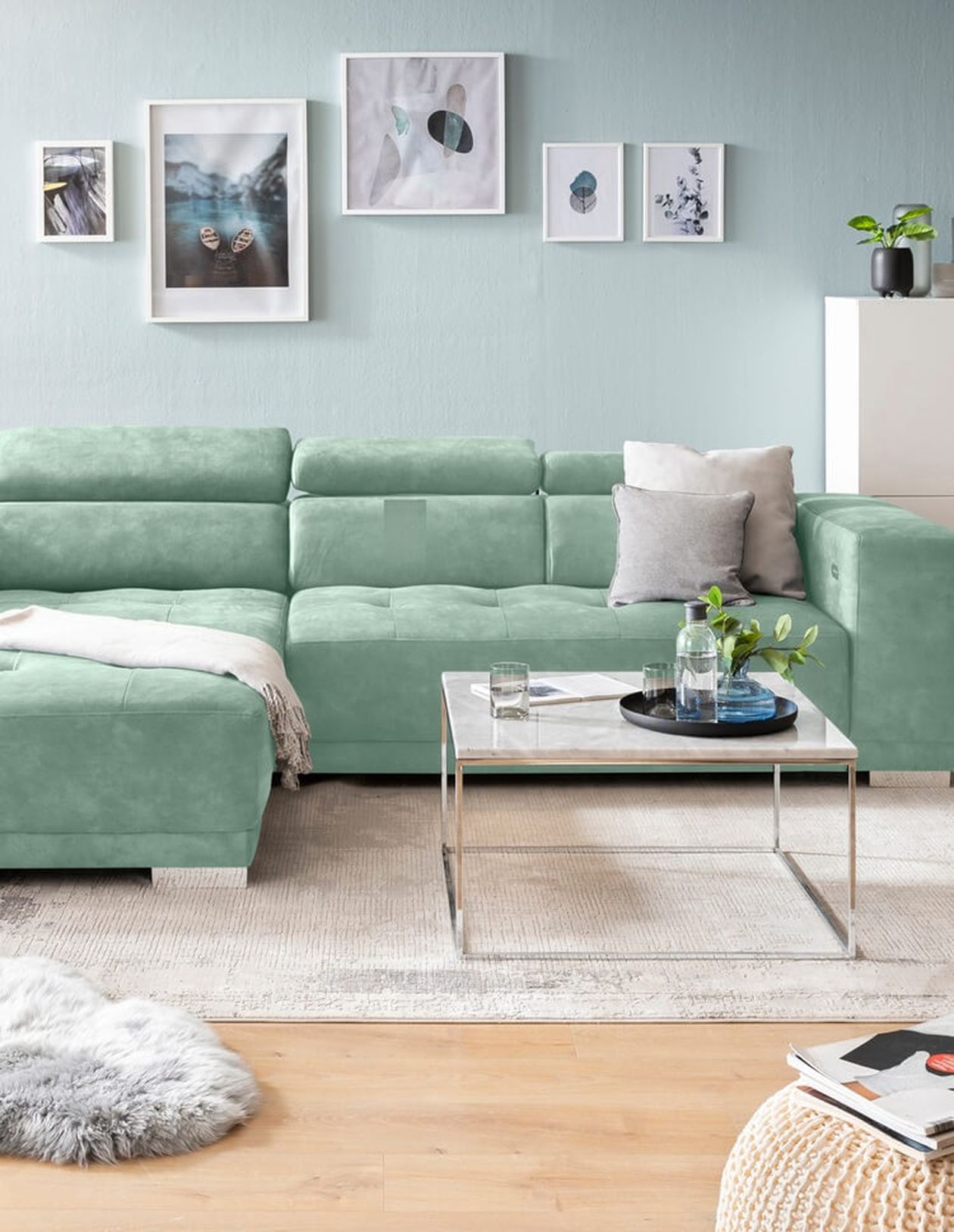 Mintgrünes Ecksofa mit Samtstoff in einem hellen Wohnzimmer mit salbeigrüner Wand. Darüber eine Bildergalerie mit maritimen Motiven. Ein Couchtisch mit Marmorplatte und Dekoelemente vervollständigen den Look.