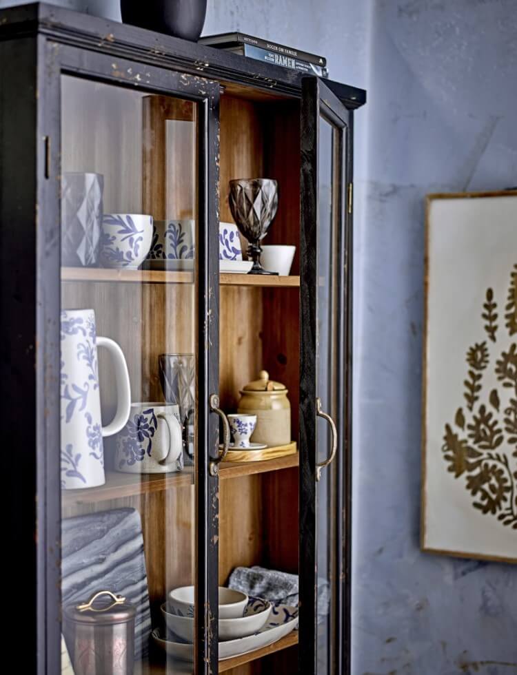 Große Vitrine aus dunklem Holz mit zwei verglasten Türen, die Blick auf Tassen, Teller und anderes Geschirr in blau-weißem Design ermöglichen. Rechts neben der Vitrine hängt ein dekoratives Bild an der Wand.
