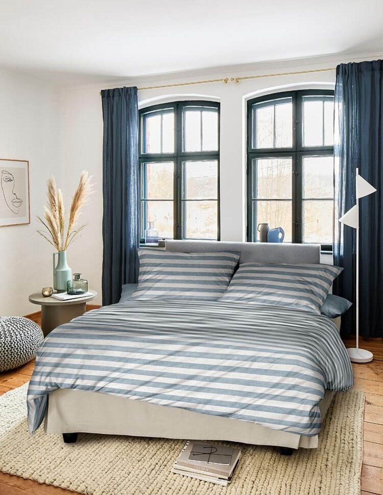 Blau-weiß gestreifte Satin-Bettwäsche auf einem Bett in einem Schlafzimmer mit Rundbogenfenstern und dunkelblauen Vorhängen. Pampasgras in einer Vase und ein Strickpouf ergänzen die Einrichtung.