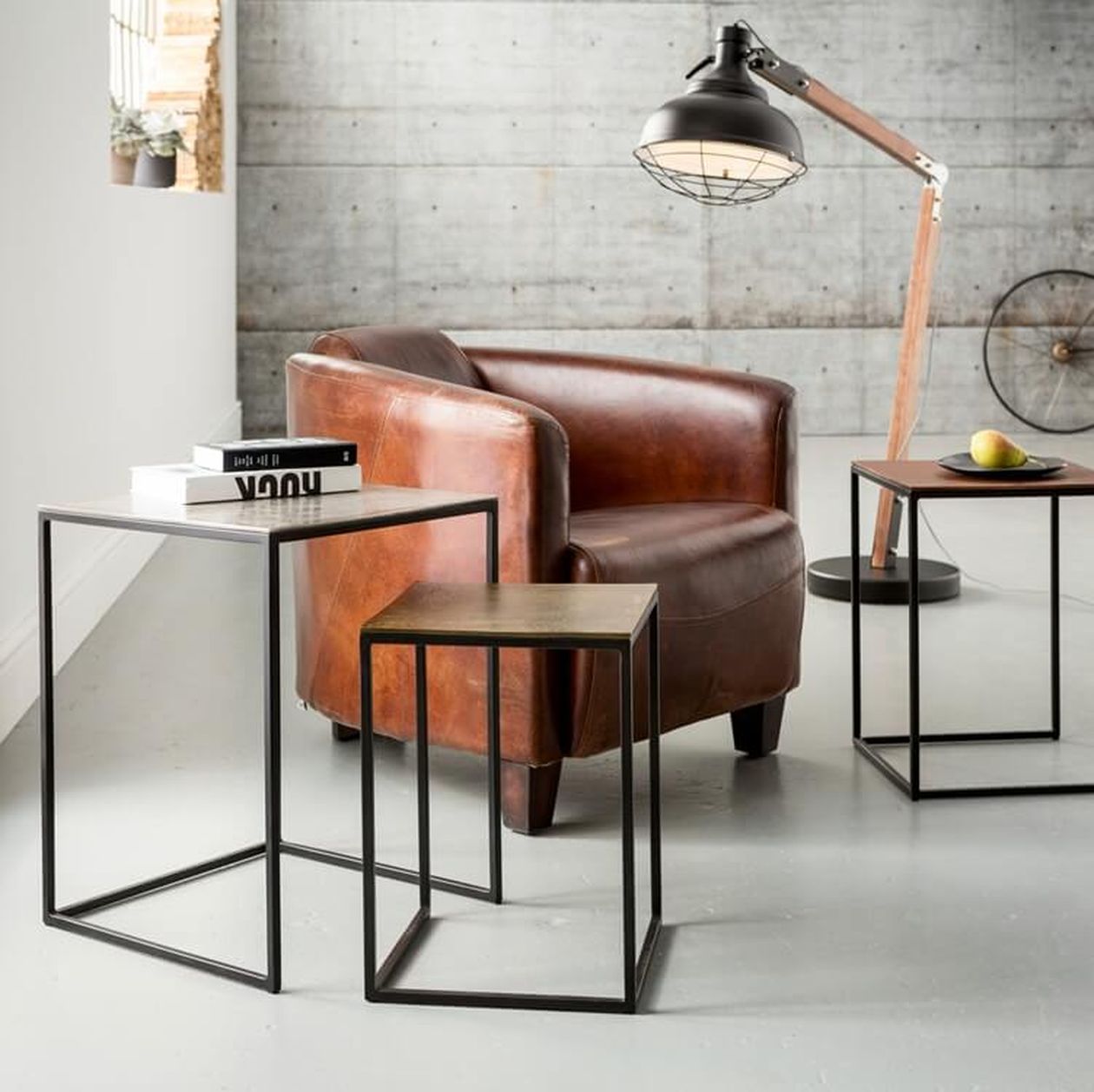 Gemütliches Zimmer mit einem braunen Ledersessel, zwei modernen Beistelltischen, Büchern, einer Lampe und einer Birne auf einem Teller vor einer Betonwand.