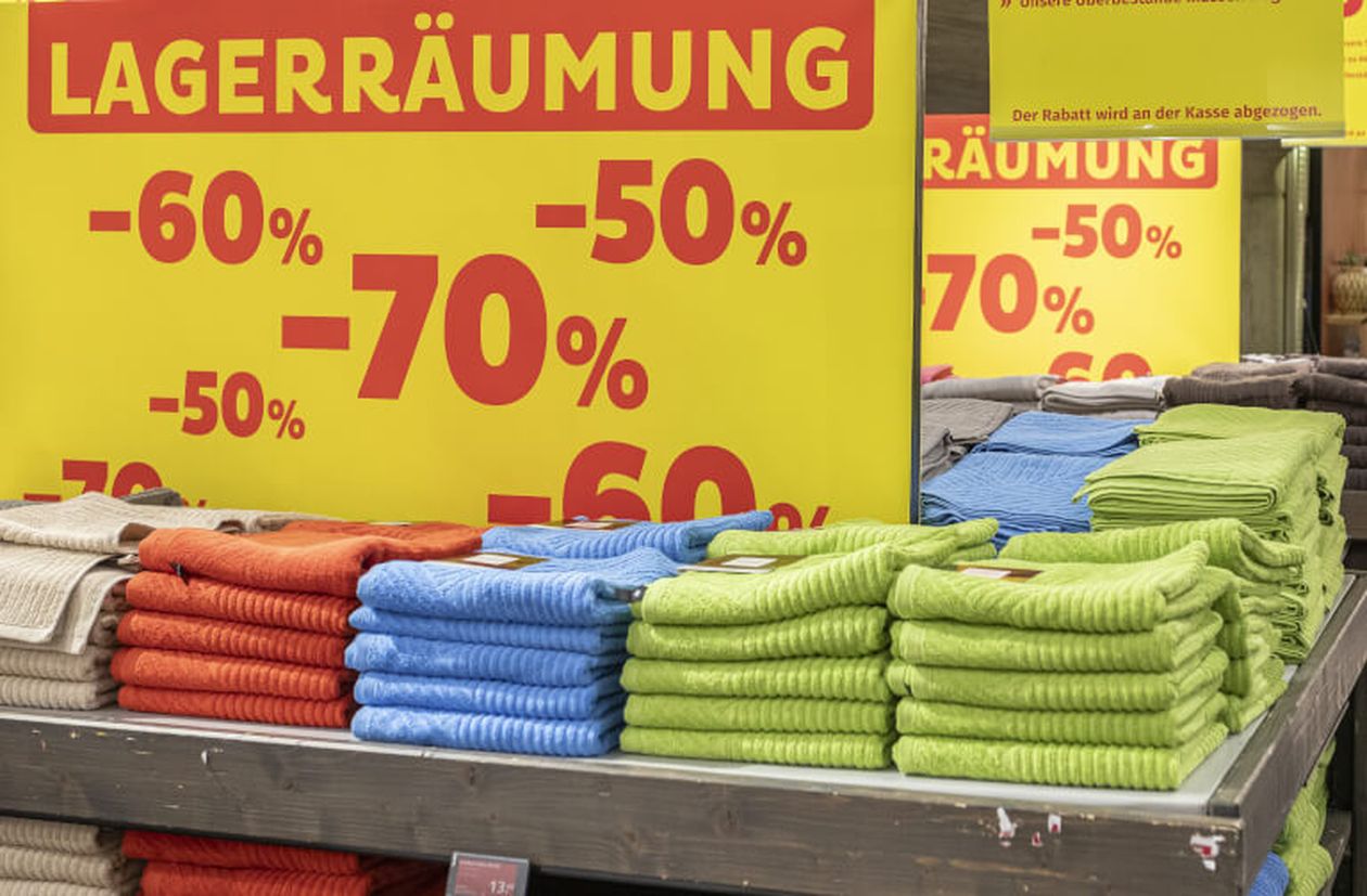 Stapel bunter Handtücher vor einem gelben Schild mit "Lagerräumung" und Rabatten von 50% bis 70% in einem Geschäft.