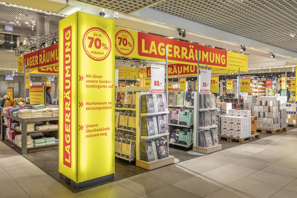 Ein Geschäft mit gelben "Lagerräumung"-Schildern bietet Rabatte bis zu 70%. Stapel von Haushaltswaren und Textilien sind ausgestellt.