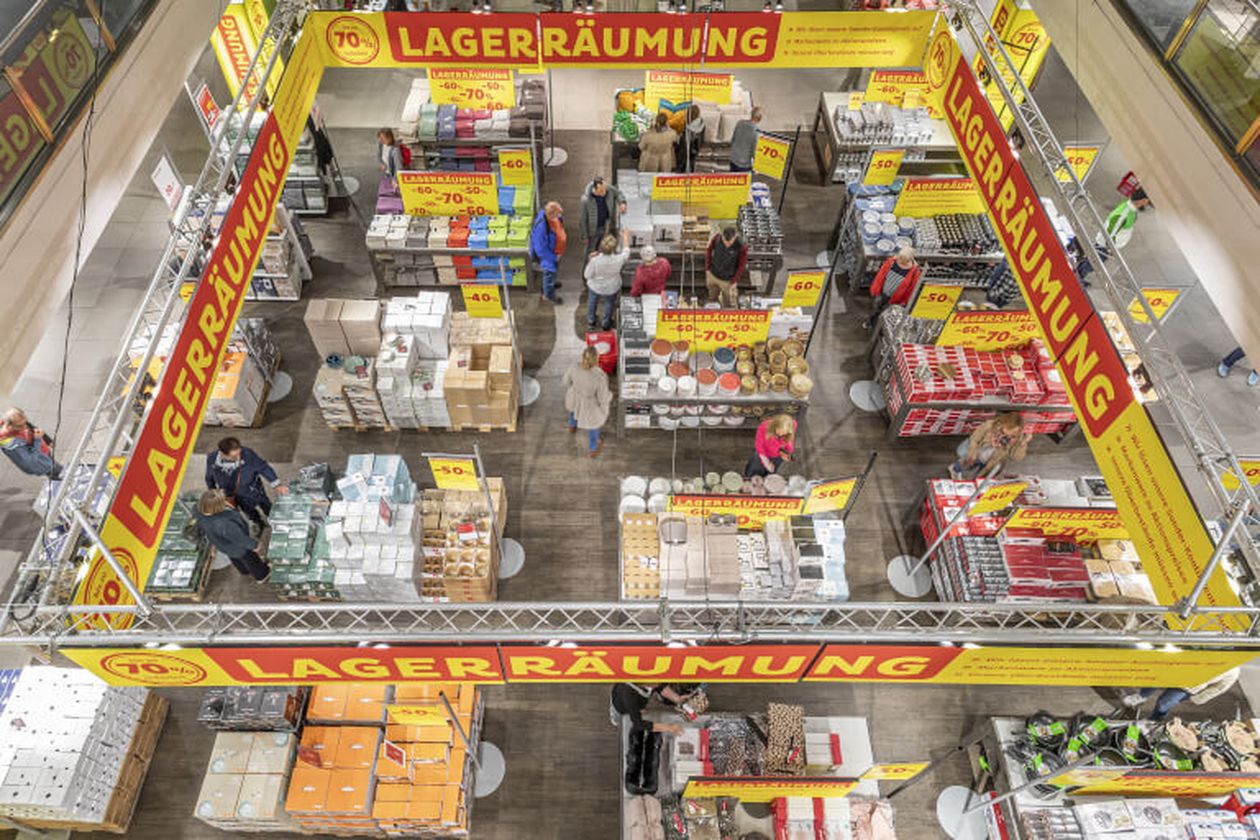 Luftaufnahme eines Geschäfts mit "Lagerräumung"-Schildern. Mehrere Regale mit Haushaltswaren und Angeboten bis zu -70% sind sichtbar. Kunden stöbern.