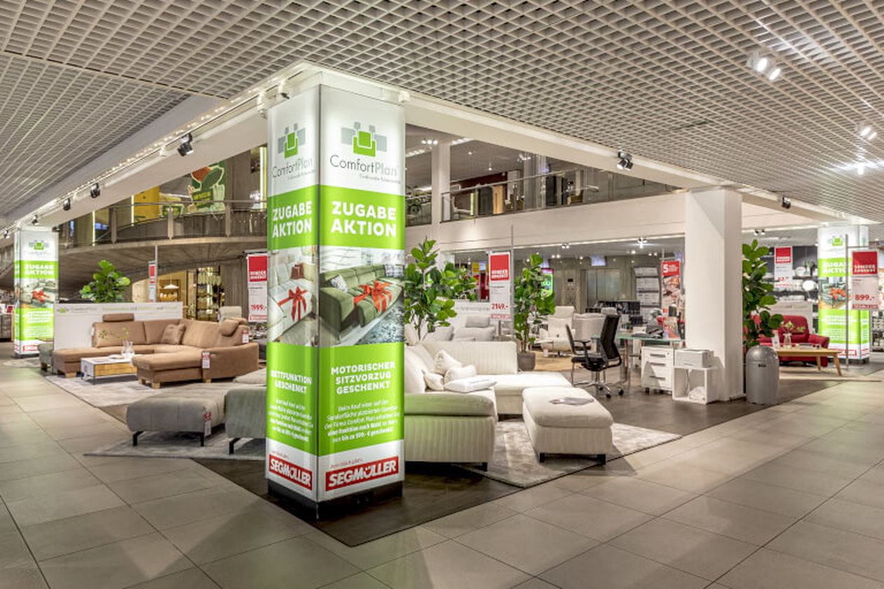 Geräumige Möbelausstellung mit Sofas, Sesseln und Dekorationen, umgeben von Werbeschildern und modernen Leuchten.