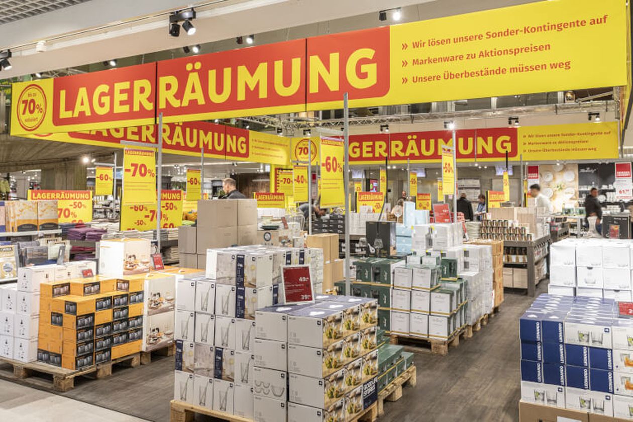 Geschäft mit großen „Lagerräumung“-Schildern, Rabatte bis 70 %, viele gestapelte Kartons mit Haushaltsartikeln, Einkaufsatmosphäre.