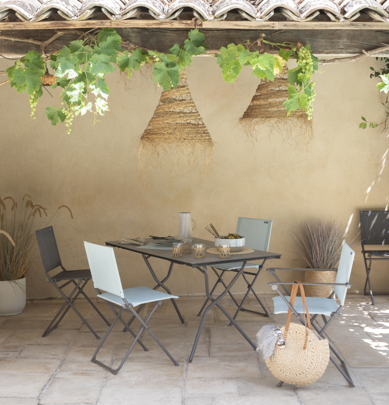 Untereinem Ziegelvordach steht die Gartenmöbel Serie Anytime von Lafuma Mobilier, bestehend aus Klapptstühlen in Türkis und Schwarz und dem passenden Gartentisch mit einer Platte in Zementoptik.