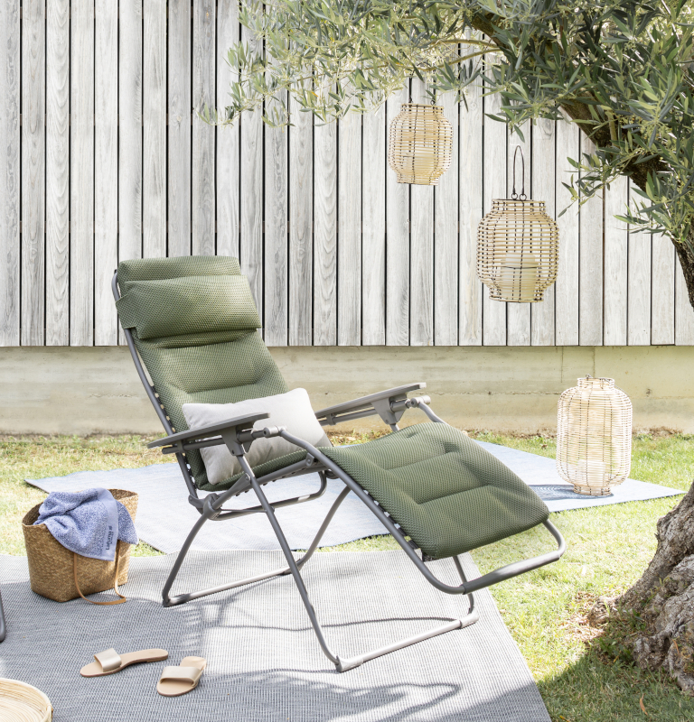 Auf einer Picknickdecke vor einem mit Laternen aus Geflecht dekoriertem Olivenbaum steht der Lafuma Mobilier Gartensessel Futura Be Comfort XL aus einem stabilen Stahlgestell und mit einem Polster in Grün.