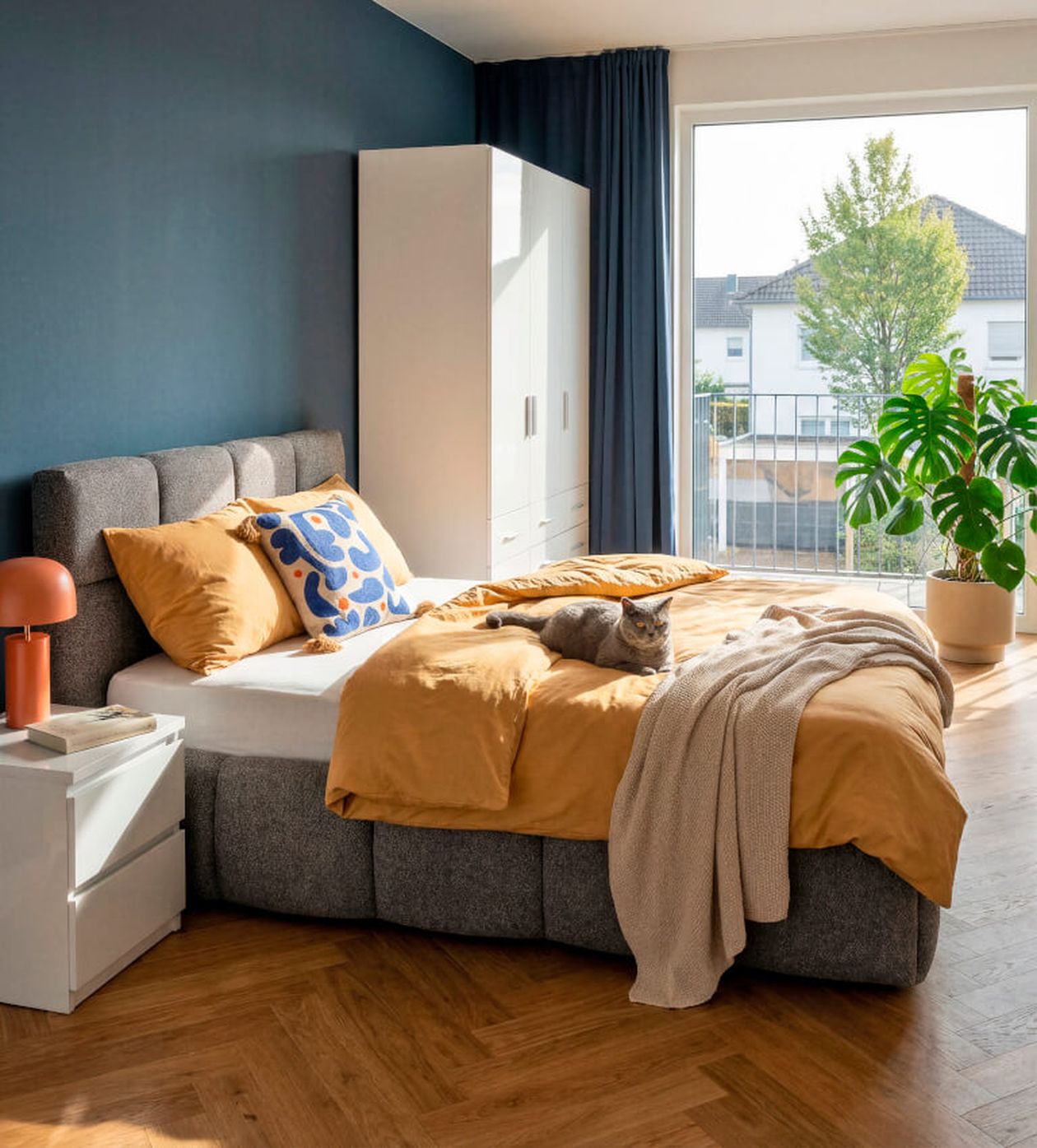 Gemütliches Schlafzimmer mit grauem Bett, senffarbener Bettwäsche und einem bunten Kissen. Eine Katze liegt auf dem Bett. Neben einem großen Fenster steht eine Pflanze, die Wände sind blau.