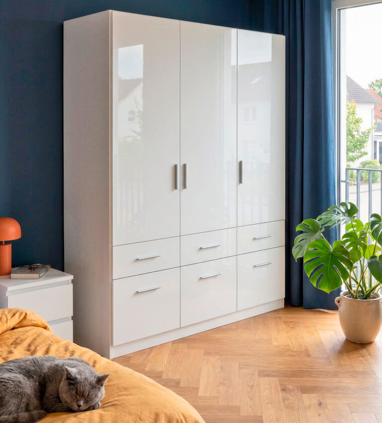 Gemütliches Schlafzimmer mit einem eleganten weißen Kleiderschrank, blauen Wänden und Vorhängen. Eine Katze schläft auf einer senffarbenen Tagesdecke neben einer Topfpflanze am Fenster.