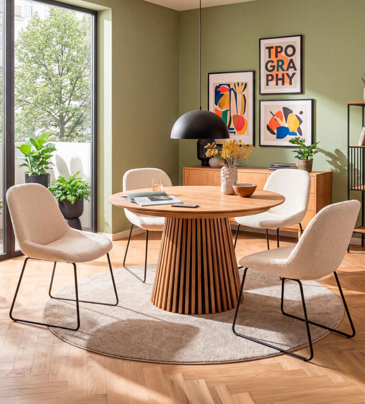 Modernes Esszimmer mit rundem Holztisch, vier beigen Stühlen, abstrakter Wandkunst, grünen Wänden und großem Fenster mit Blick auf Pflanzen.