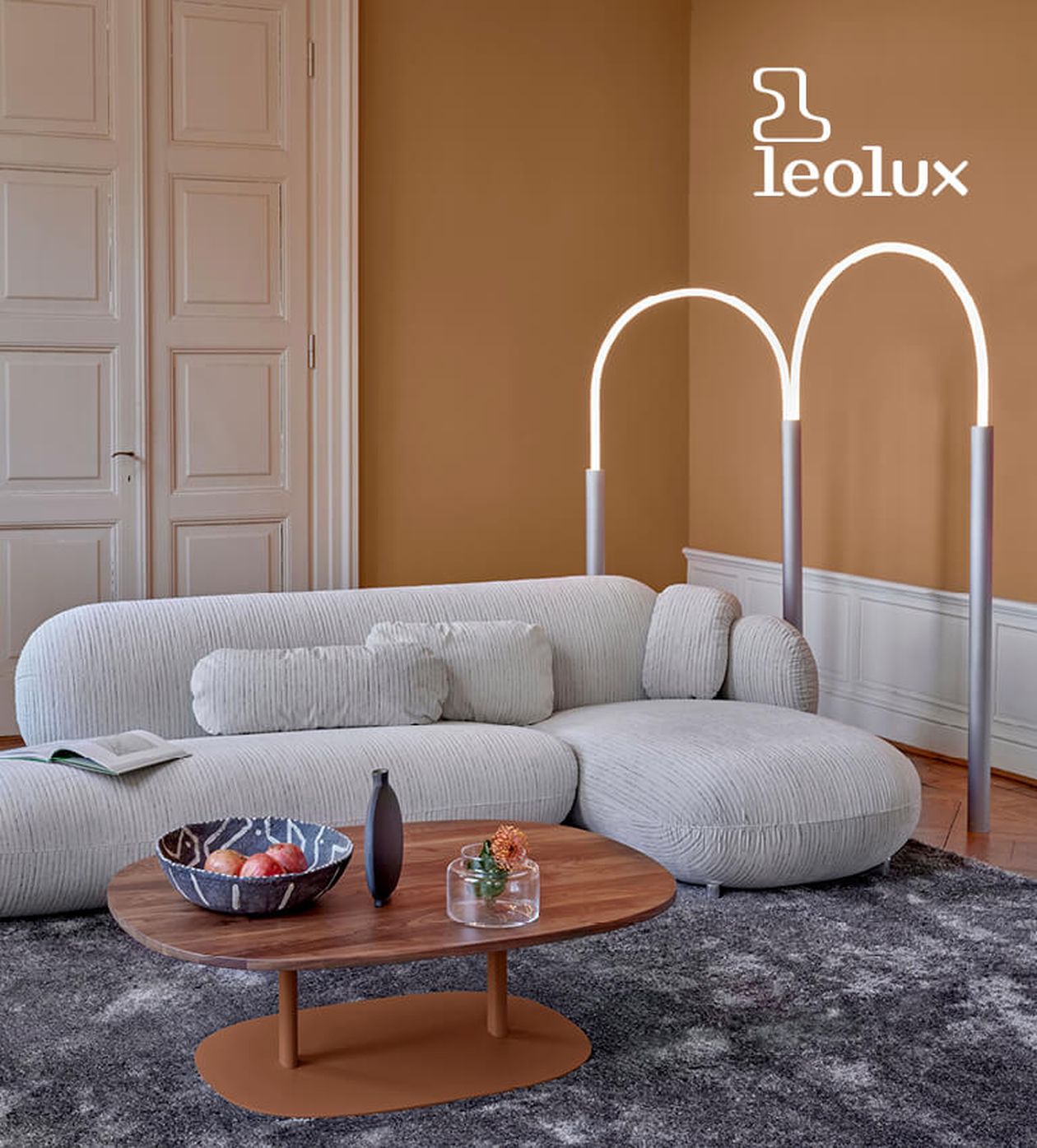 Modernes Wohnzimmer mit geschwungenem weißen Sofa, Couchtisch aus Holz und Bogenstehlampe. Ein strukturierter Teppich unterstreicht die warmen braunen Wände.