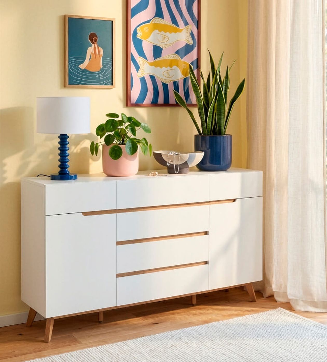 Modernes weißes Sideboard mit Pflanzen und einer Lampe, verziert mit farbenfrohen Kunstwerken an einer sanft beleuchteten gelben Wand, in der Nähe von transparenten Vorhängen.