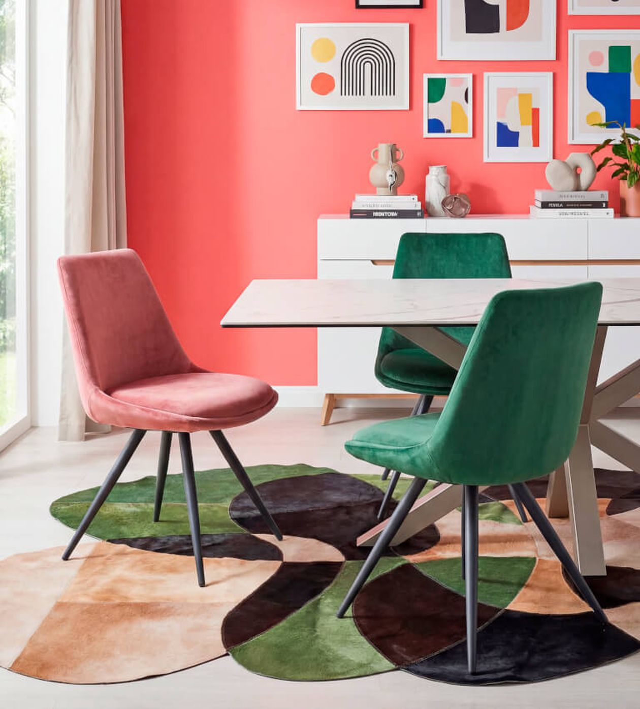 Modernes Esszimmer mit rosa und grünen Stühlen, abstrakter Kunst an einer korallenroten Wand und einem geometrischen Teppich. Weiße Kommode und Dekoration im Hintergrund.