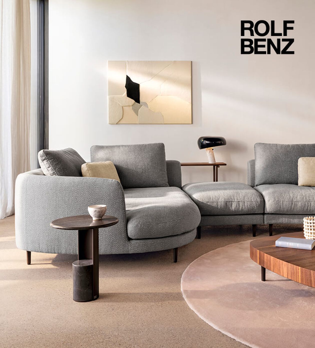 Modernes Wohnzimmer mit grauem, geschwungenem Sofa, abstrakter Wandkunst, runden Couchtischen und einer schwarzen Lampe. Logo „Rolf Benz“ in der oberen rechten Ecke.