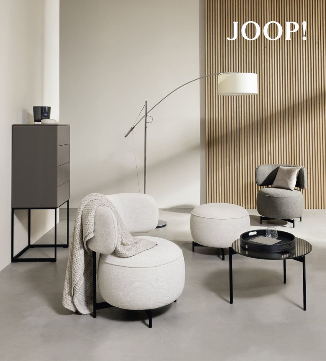 Moderne Wohnzimmermöbel mit cremefarbenen Sesseln, grauer Kommode, runder Beistelltisch und Stehlampe vor holzverkleideter Wand.