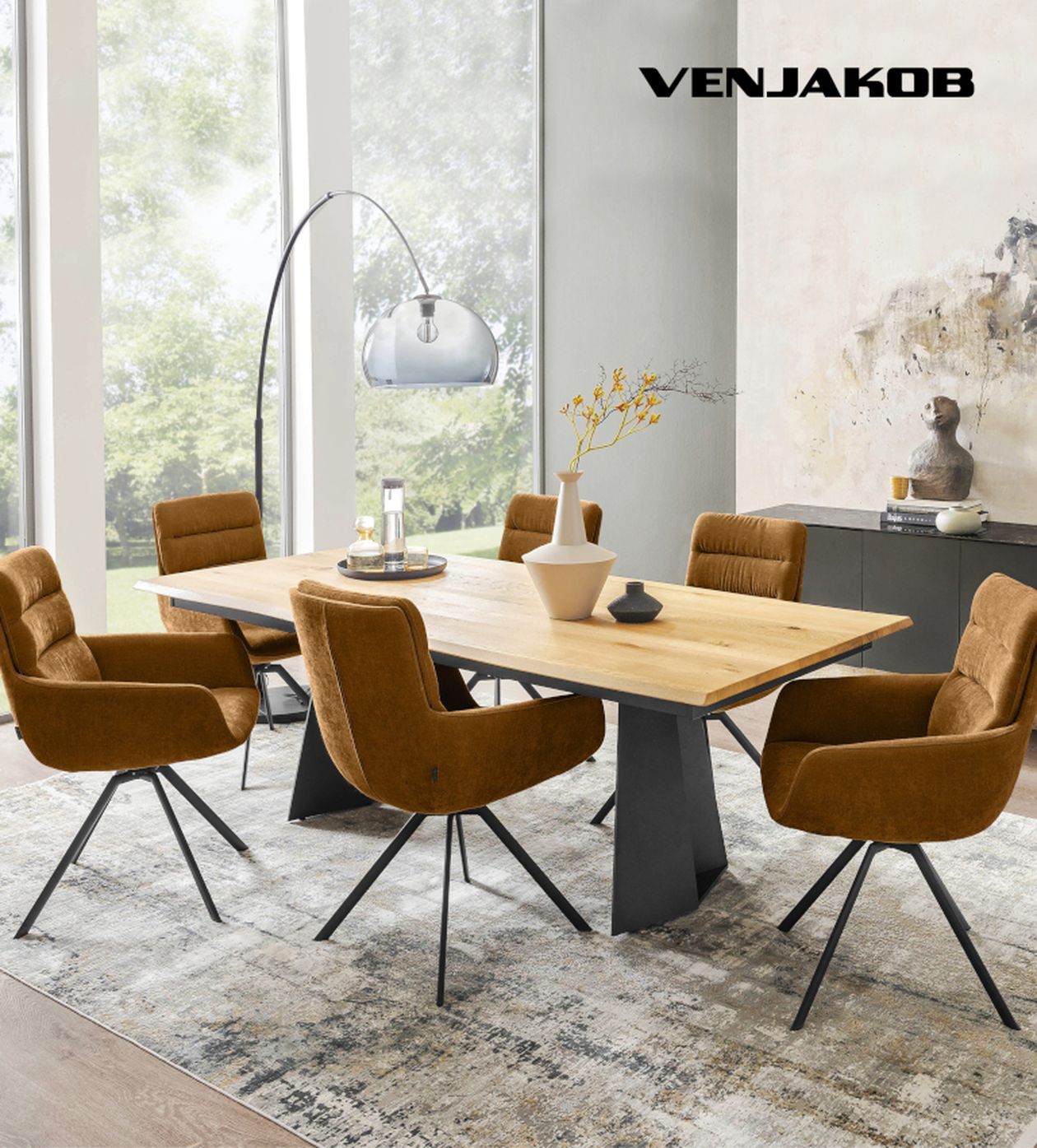 Esszimmer mit Holztisch, sechs braunen Polsterstühlen, einer Stehlampe und Dekorationen, große Fenster im Hintergrund, helle, moderne Atmosphäre.