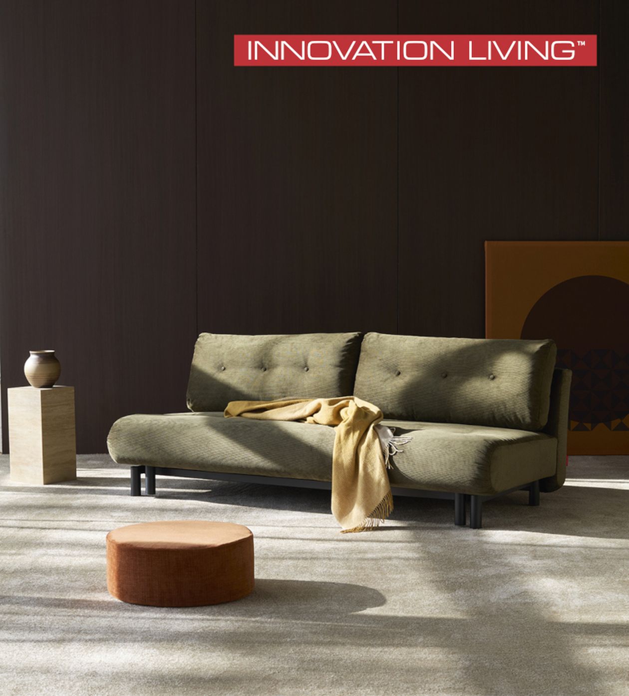 Grünes Sofa mit gelber Decke in modernem Wohnzimmer, brauner Hocker und Vase auf Sockel, Marke "Innovation Living" oben.