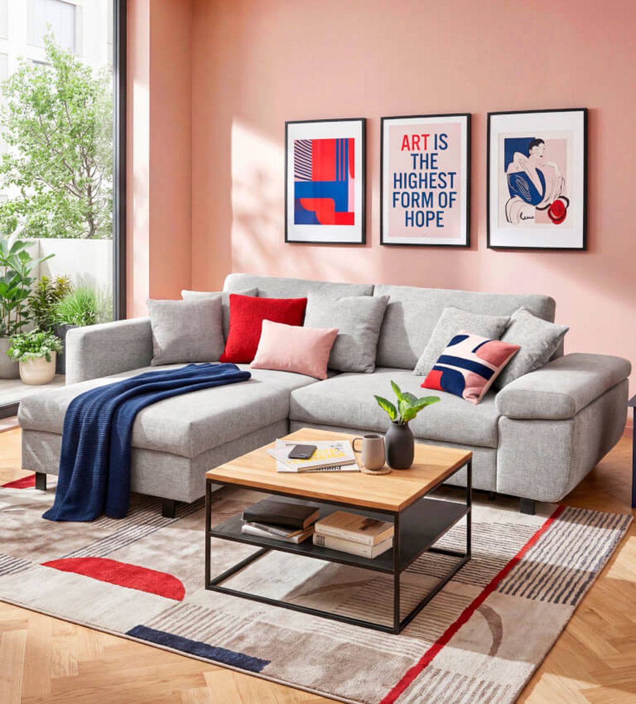 Ein modernes Wohnzimmer mit grauem Ecksofa, farbenfrohen Kissen, einem Couchtisch aus Holz und abstrakter Kunst an rosa Wänden. Großes Fenster mit Blick ins Grüne.