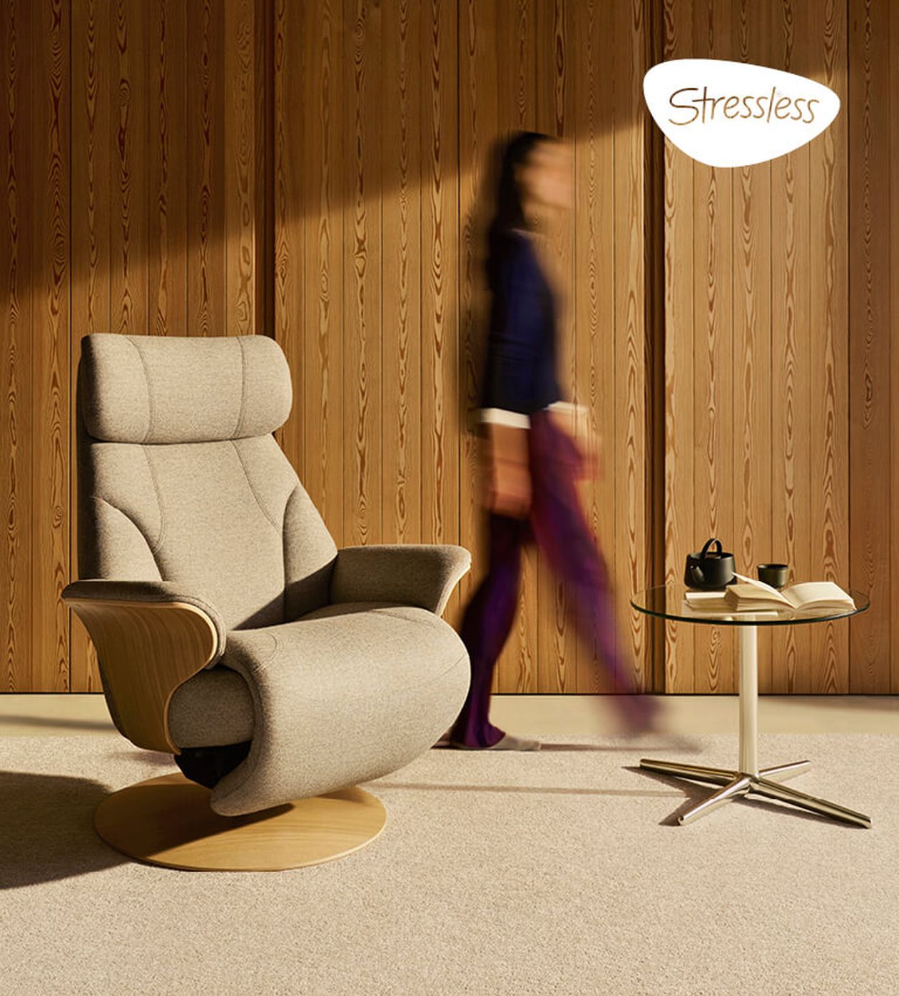 Ein beigefarbener Sessel mit Holzelementen neben einem Glastisch mit Teekanne und Buch. Eine verschwommene Person geht an einer Holzvertäfelung vorbei. „Stressless“-Logo in der Ecke.