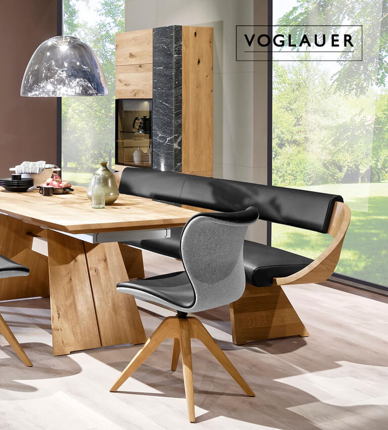 Modernes Esszimmer mit Holztisch, gepolsterter Bank, stilvollen Stühlen und großem Fenster. Marke: „Voglauer“.