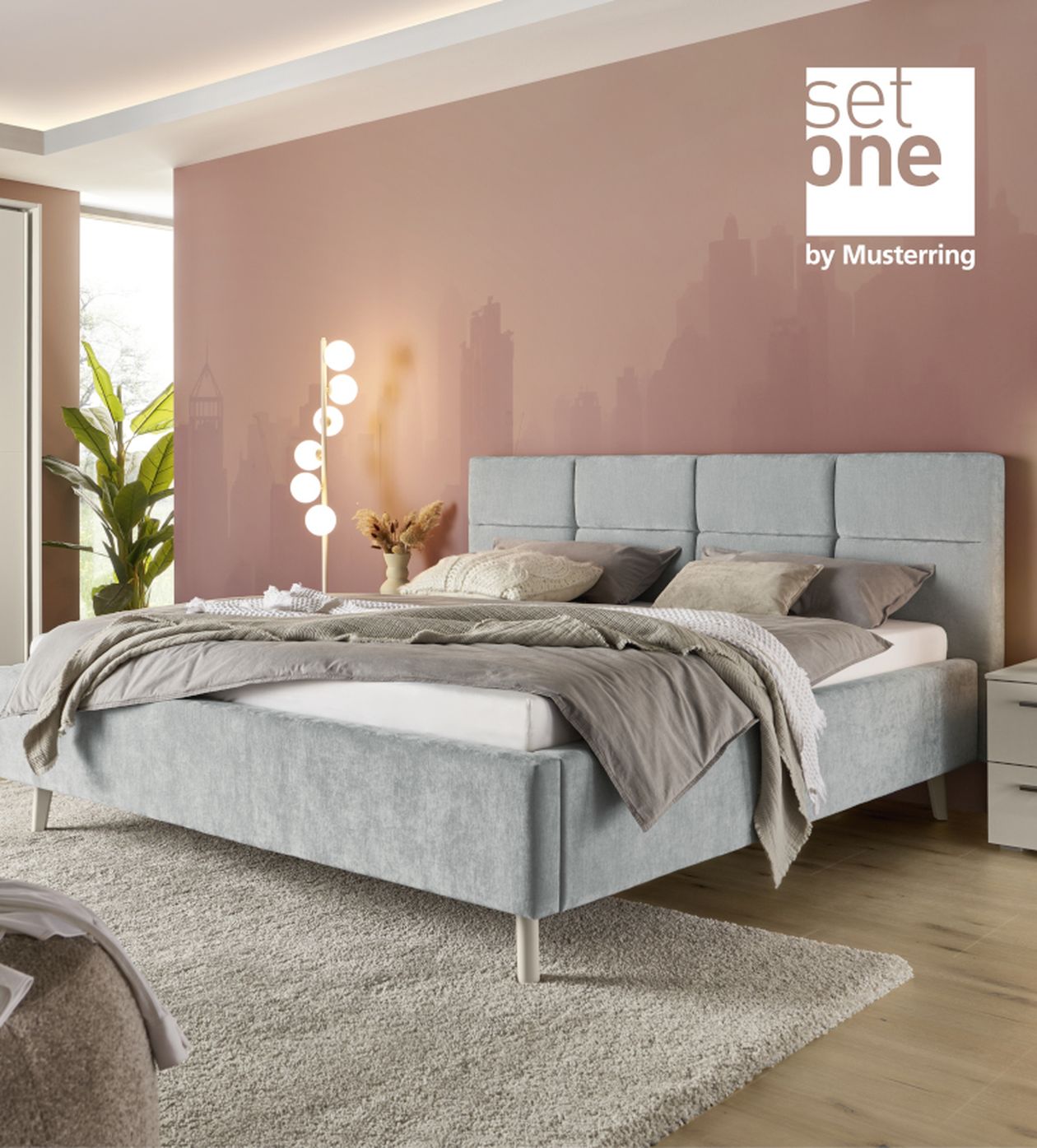 Modernes Schlafzimmer mit grauem Polsterbett, pastellrosa Wand, Nachttisch, Stehlampe und Pflanzen. Teppich bedeckt den Holzboden.