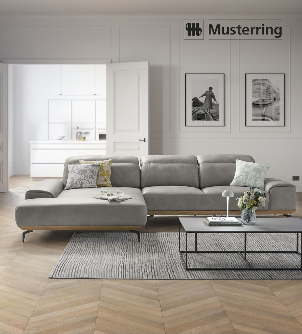 Graues Ecksofa mit bunten Kissen in modernem Wohnzimmer, Couchtisch mit Lampe und Blumen, Holzboden, offene Tür zur hellen Küche.