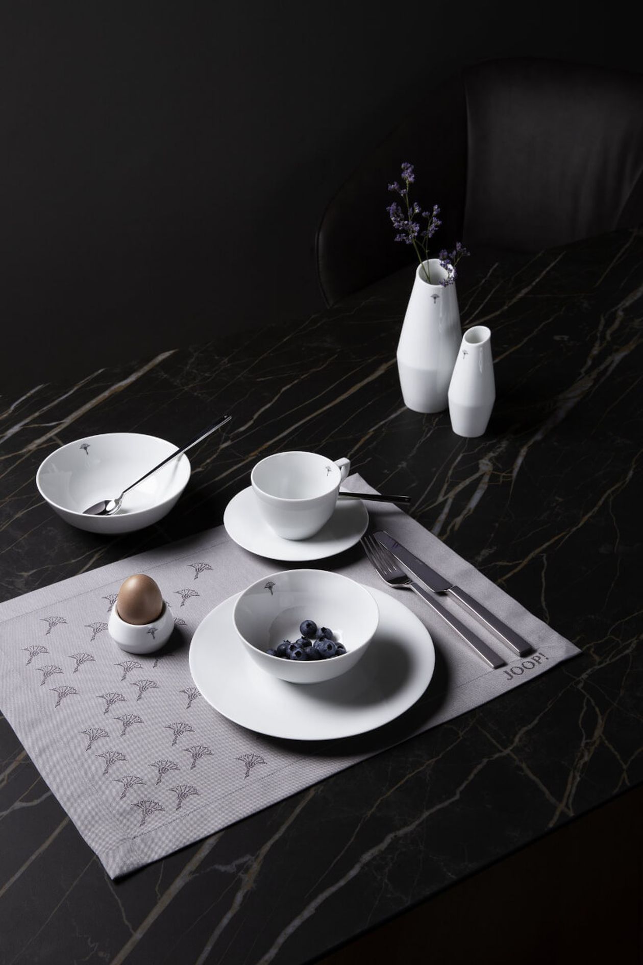 Eine elegante Tischdekoration mit weißem Geschirr auf einer gemusterten Tischset, garniert mit Heidelbeeren, einem Ei und minimalistischen Vasen mit Blumen.