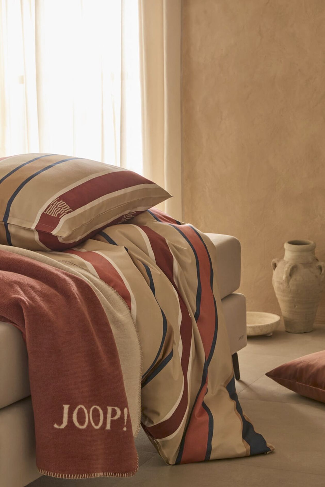 Ein beiges Sofa mit gestreifter Bettwäsche in Rot, Beige und Blau. Darauf liegen ein Kissen und eine Decke mit der Aufschrift „JOOP!“. Im Hintergrund steht eine Tonvase.