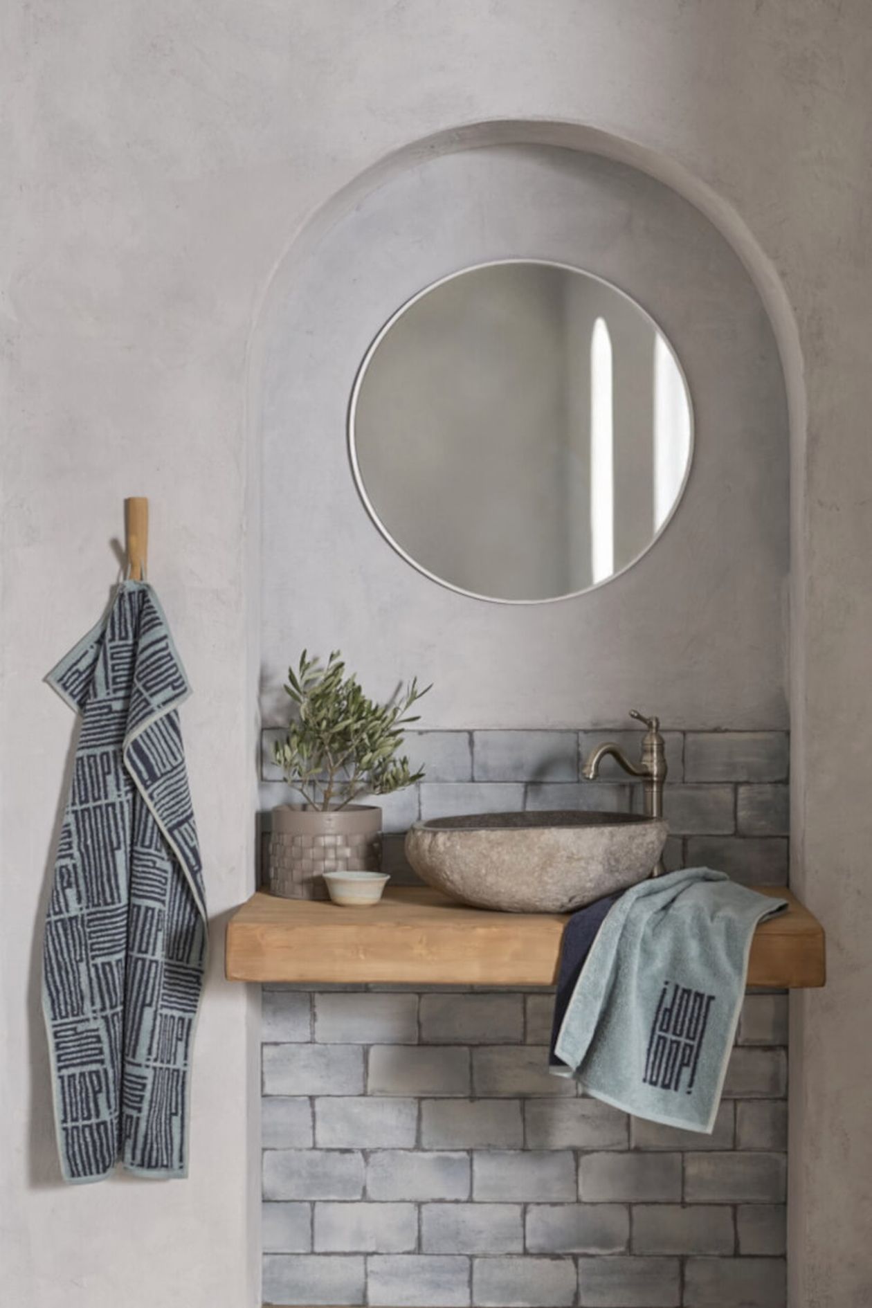 Badezimmer mit einem Waschbecken aus Stein, einem runden Spiegel und einem Holzregal. In der Nähe hängen Handtücher; ein kleiner Olivenbaum sorgt für einen Hauch von Grün.