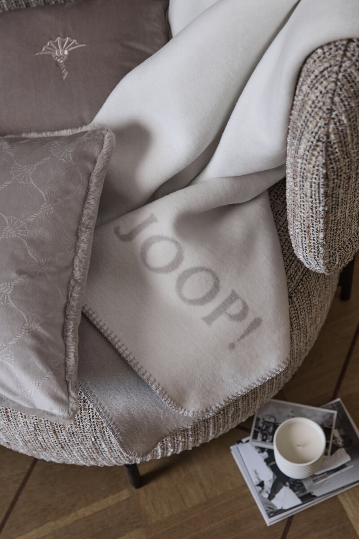 Ein gemütlicher Sessel mit einer weichen weißen „JOOP!“-Decke und grauen Kissen; daneben ein Stapel Zeitschriften und eine brennende Kerze auf dem Boden.