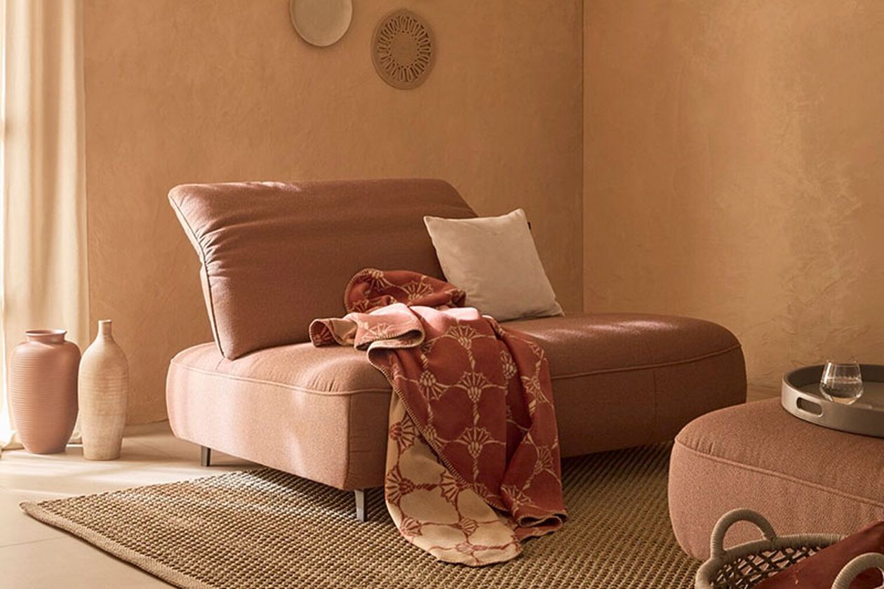 Gemütliches Wohnzimmer mit einer rosa Chaiselongue, einer gemusterten Decke und einem Kissen auf einem strukturierten Teppich. Beige Wände und dekorative Vasen unterstreichen die warme Atmosphäre.