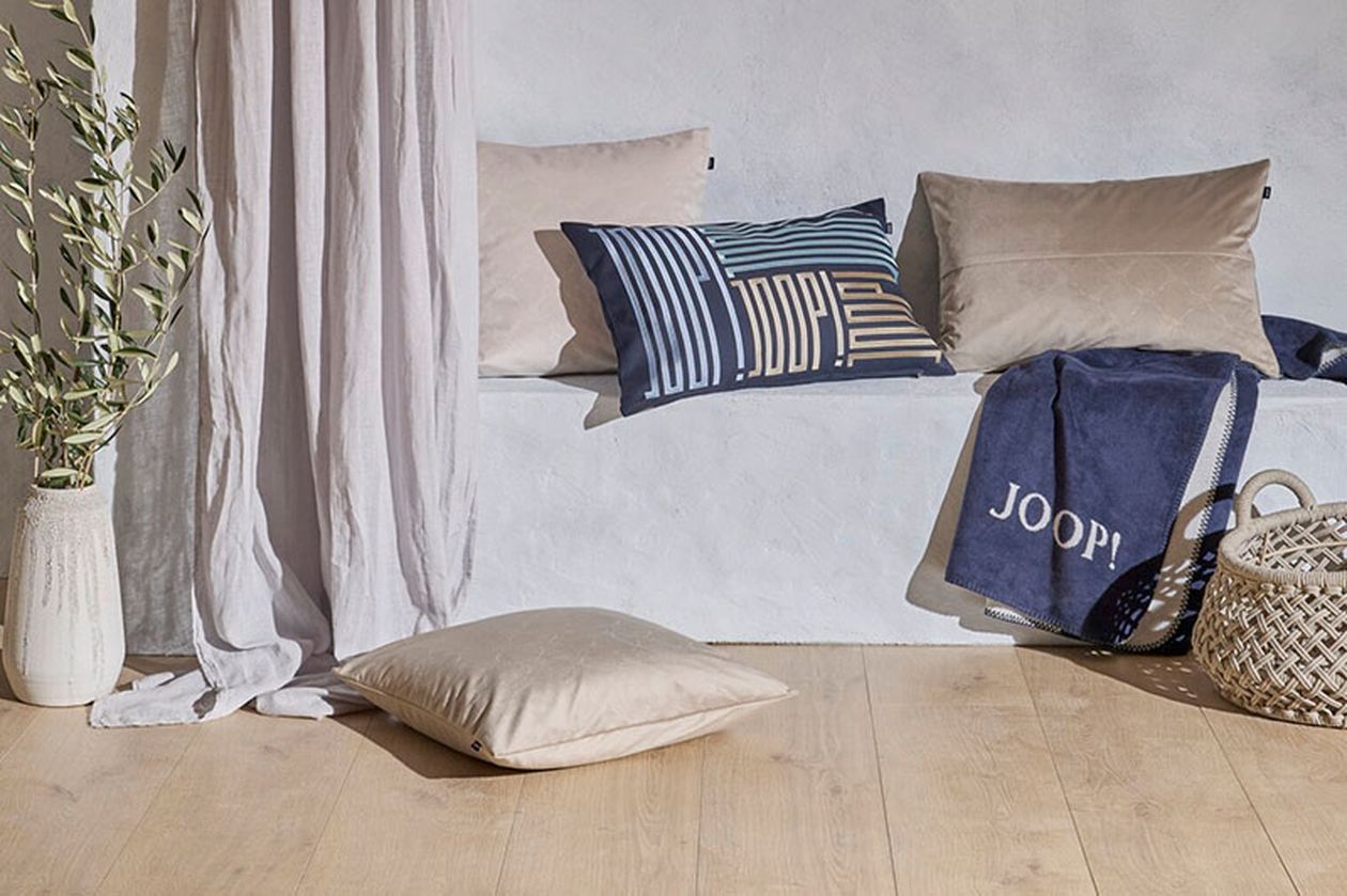 Gemütliche Sitzecke mit beigefarbenen Kissen, einem blau gemusterten Kissen, einer blauen Überwurfdecke und einem geflochtenen Korb auf einem Holzboden.