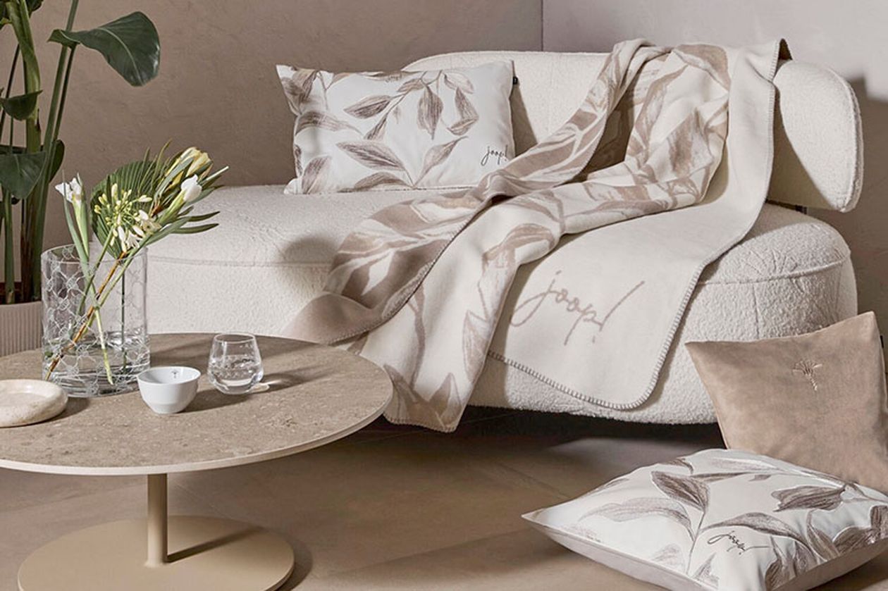 Gemütliches Wohnzimmer mit weißem Sofa, geblümtem Kissen, passender Decke, rundem beigefarbenem Tisch, Glasvasen und Zierpflanzen.