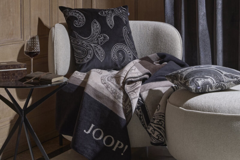 Produktbild: Kissenbezüge der Marke JOOP! auf einem Sofa in einem dunkel gehaltenen Wohnzimmer.