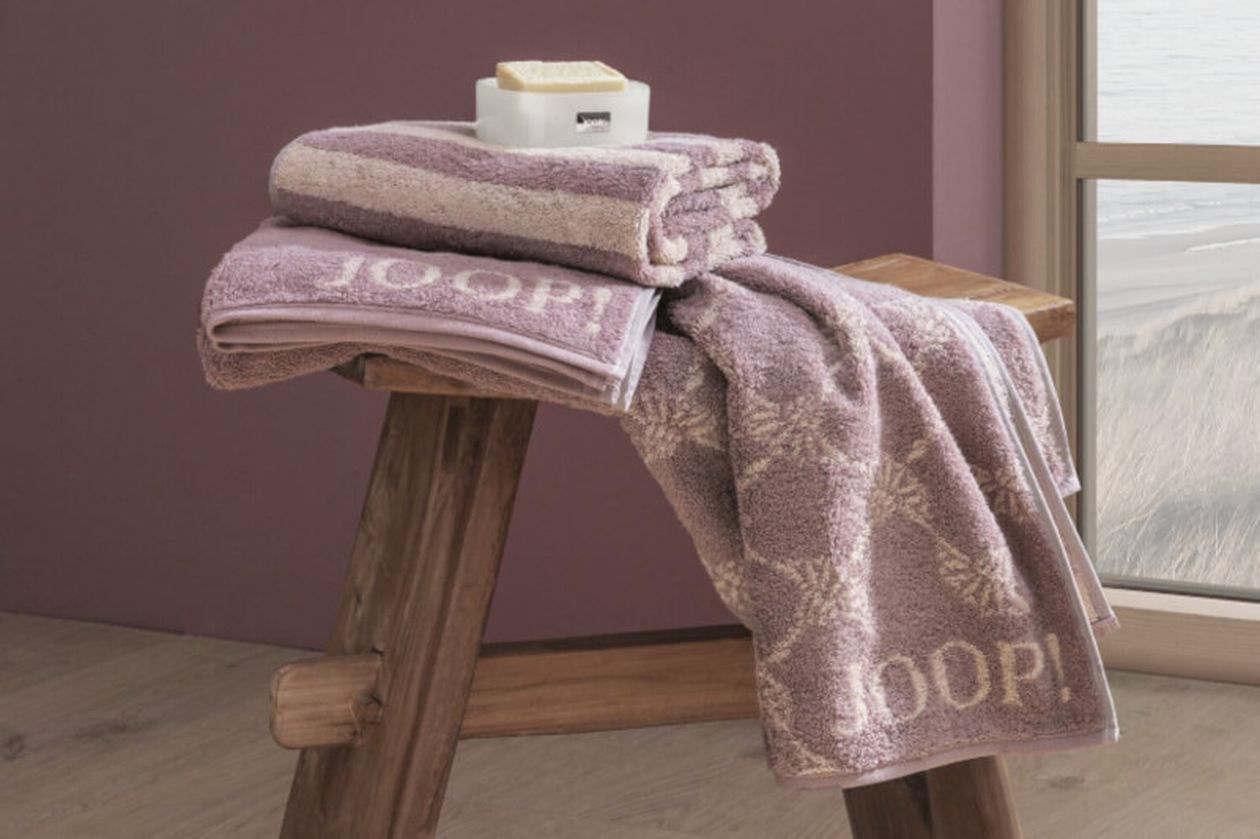 Produktbild: Handtücher und Seiftücher der Marke JOOP! auf einem hölzernen Hocker in einem rosafarbigen Badezimmer.